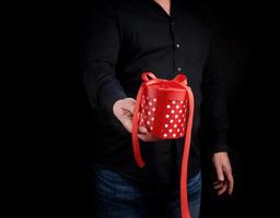 volwassen Mens in een zwart overhemd houdt in zijn hand- een ronde rood doos van polka dots met een boog Aan een zwart achtergrond foto