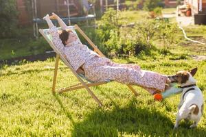 vrouw in pyjama- resting in stoel Aan groen gazon Aan zonnig zomer dag en hond foto