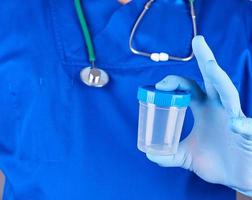 de dokter in blauw uniform en latex handschoenen is Holding een leeg plastic houder voor nemen urine monsters foto