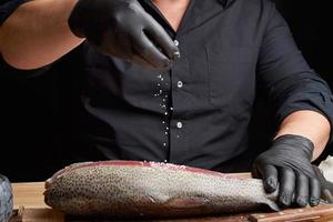 chef in een zwart overhemd en zwart latex handschoenen bereidt zich voor Zalm filet Aan een houten snijdend bord foto