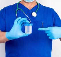 dokter in blauw uniform en latex handschoenen is Holding een leeg plastic houder voor nemen urine monsters foto