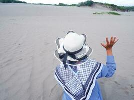 terug van de vrouw in de hoed wie was zittend Aan de wit strand zand terwijl verhogen haar hand- Hoi foto