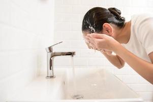 mooi Aziatisch vrouw is het wassen haar gezicht in badkamer Bij huis foto