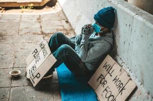 man zit naast de straat met een medisch masker met een daklozenbericht foto