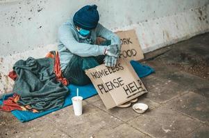 man zit naast de straat met een medisch masker met een daklozenbericht foto