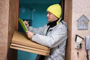 levering Mens met pizza karton dozen. koerier in groen kleur hoed Holding een klembord terwijl staand tegen deur van woon- huis foto