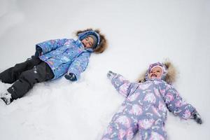 twee zussen maken sneeuw engel terwijl aan het liegen Aan sneeuw. foto