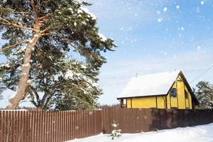 een geel knus huis in de sneeuw in winter in de dorp is omringd door pijnboom bomen. met sneeuw bedekt dak, verwarming en ventilatie pijpen, trapezium ramen foto