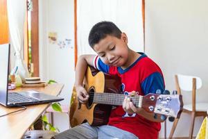 de verhaal van een jongen aan het kijken een notitieboekje computer terwijl voorbereidingen treffen naar praktijk spelen gitaar Bij huis. jongens nemen klassiek gitaar lessen online. foto