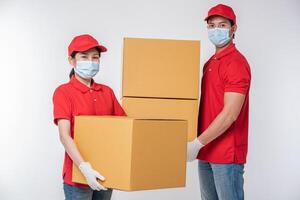 beeld van een bewust jong levering Mens in rood pet blanco t-shirt uniform gezicht masker handschoenen staand met leeg bruin karton doos geïsoleerd Aan licht grijs achtergrond studio foto