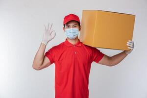 beeld van een bewust jong levering Mens in rood pet blanco t-shirt uniform gezicht masker handschoenen staand met leeg bruin karton doos geïsoleerd Aan licht grijs achtergrond studio foto