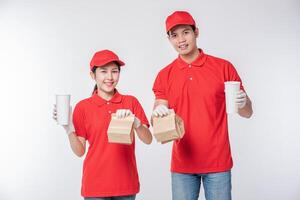 beeld van een gelukkig jong levering Mens in rood pet blanco t-shirt uniform gezicht masker handschoenen staand met leeg bruin ambacht papier pakket geïsoleerd Aan licht grijs achtergrond studio foto