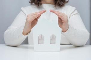 vrouw handen gevouwen naar elk andere over- een houten miniatuur model- huis Aan een wit achtergrond. echt landgoed verzekering concept, milieu bescherming foto