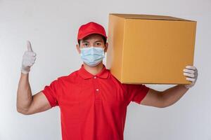 beeld van een bewust jong levering Mens in rood pet blanco t-shirt uniform gezicht masker handschoenen staand met leeg bruin karton doos geïsoleerd Aan licht grijs achtergrond studio foto