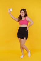 portret van glimlachen jong vrouw selfie met mobiel telefoon in haar handen terwijl staand geïsoleerd over- geel achtergrond foto