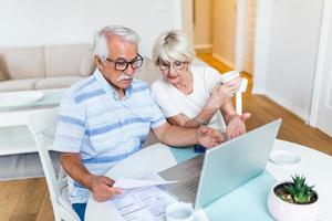 gelukkig oud man en vrouw houden papieren gebruik makend van laptop voor online bankieren. hebben een kop van thee foto