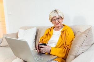 senior vrouw besteedt huis vrije tijd met een laptop. grijnzend ouderen vrouw zit Bij een knus bank, Holding kop van koffie of thee. web browsen, aan het kijken TV serie, boodschappen doen in netwerk,video telefoontje foto