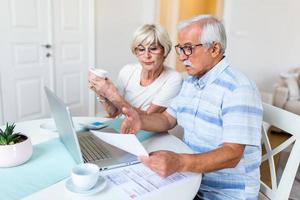 gelukkig oud man en vrouw houden papieren gebruik makend van laptop voor online bankieren. drinken koffie foto