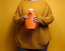 vrouw in een gebreid trui houdt een oranje plastic fles met vloeistof het wassen gel. wasserij en klusjes, geel achtergrond foto