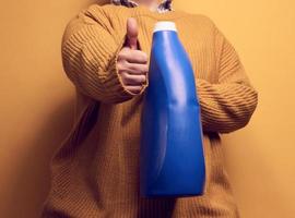 vrouw in een gebreid trui houdt blauw plastic flessen met vloeistof het wassen gel. wasserij en klusjes, geel achtergrond foto