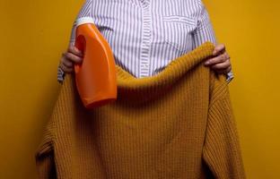 een vrouw in een wit gestreept overhemd houdt een oranje plastic fles met vloeistof het wassen gel en een gebreid trui. wasserij en huishouden klusjes, schoon gewassen items foto