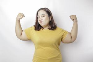 opgewonden Aziatisch groot formaat vrouw vervelend een geel t-shirt tonen sterk gebaar door hijs- haar armen en spieren glimlachen trots foto