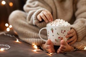 kind kaal voeten en handen houden kop heet cacao marshmallows dichtbij omhoog slinger lichten.flaffy wazig warm gebreid beige trui. Kerstmis concept, vakantie.blij nieuw jaar.kind meisje zittend Aan sofa huis foto