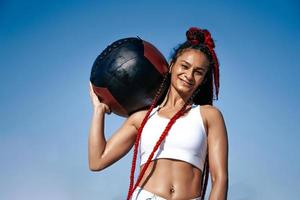 houdt Aan schouder med bal. atletisch vrouw met med bal. sterkte en motivatie.foto van sportief vrouw in modieus sportkleding achtergrond lucht foto