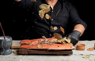 volwassen Mens in een zwart overhemd giet wit grof zout en een droog baai blad Aan een vers Zalm filet foto