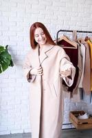 jong mooi vrouw vervelend beige pastel jas, staand in de buurt kleren rek. garderobe verandering foto