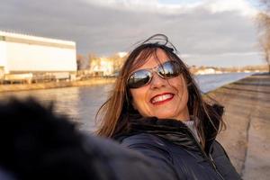 midden- oud vrouw vervelend winter kleren nemen een selfie door een rivier- foto