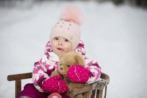 een weinig meisje zit in een houten slee met een teddy beer in winter. foto