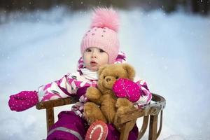 een weinig meisje zit in een houten slee met een teddy beer in winter. kind Aan een winter wandelen. foto