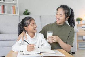 Aziatisch jong weinig meisje leren Bij huis. Doen huiswerk met soort moeder helpen, aanmoedigen voor examen. mam voorbij gaan aan Aan een glas van melk naar dochter. meisje gelukkig thuisschool. mam onderwijzen en advies onderwijs samen. foto