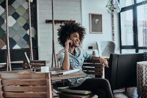 altijd in tintje met klanten. mooi jong Afrikaanse vrouw pratend Aan slim telefoon en glimlachen terwijl zittend Bij haar werken plaats foto