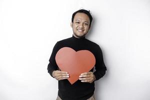 een portret van een gelukkig Aziatisch Mens vervelend een zwart shirt, Holding een rood hartvormig papier geïsoleerd door wit achtergrond foto