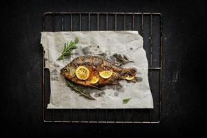 gebakken vis gekookt Aan perkament papier met kruiden, citroen en rozemarijn. barbecue rooster Aan een donker achtergrond. foto