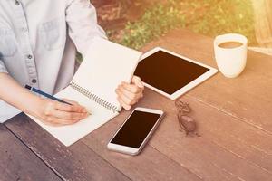 vrouw hand- schrijven notitieboekje en telefoon, tablet Aan tafel in tuin Bij koffie winkel met wijnoogst afgezwakt. foto