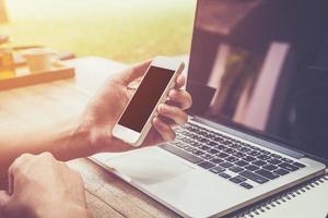 jong bedrijf Mens hand- Holding telefoon en gebruik makend van laptop computer in koffie winkel met wijnoogst afgezwakt. foto