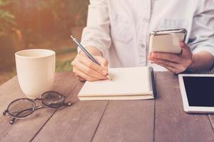 dichtbij omhoog hand- vrouw schrijven notitieboekje en Holding telefoon in koffie winkel met wijnoogst afgezwakt. foto