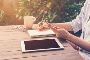 dichtbij omhoog hand- vrouw schrijven notitieboekje en Holding telefoon in koffie winkel met wijnoogst afgezwakt. foto