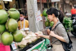 gelukkig jong Aziatisch vrouw rugzak reiziger staan in voorkant van kokosnoot sap winkel Bij China stad- straat voedsel markt in Bangkok, Thailand. reiziger controle uit kant straten. foto