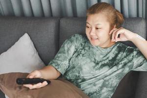 Aziatisch huisvrouw zitten in sofa en Holding afgelegen controle aan het kijken televisie met ontspannende actie. hobby en vrij tijd leven stijl concept. foto