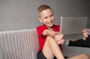 een gelukkig jongen in een rood t-shirt is zittend Aan de bankstel met een tablet en lachend foto