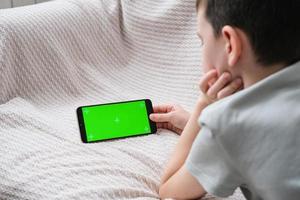 de jongen looks Bij de scherm van de telefoon met een chroom sleutel binnenshuis. een onherkenbaar kind toepassingen een smartphone met een groen scherm naar kijk maar een video. foto
