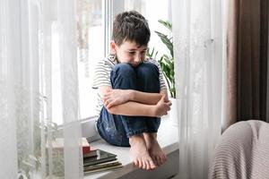 een verdrietig jongen zit Aan de vensterbank knuffelen zijn knieën. slecht stemming, depressie. de jongen is verdrietig alleen Bij huis foto