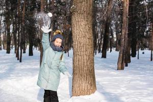 een vrolijk meisje in warm kleren gooit een sneeuwbal in de winter Woud. wandelen buitenshuis. foto