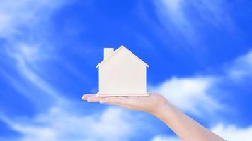 vrouw hand- Holding klein huis model- houten met wolk blauw lucht achtergrond. familie leven en bedrijf echt landgoed concept, eco huis foto