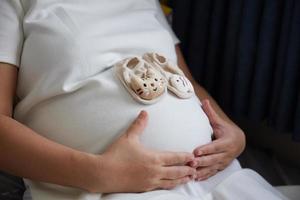 foto van gelukkig Aziatisch zwanger vrouw zittend in bed en aanraken haar buik Bij huis met klein schoenen voor de ongeboren baby in de buik