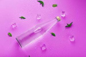 een fles met een ijs drinken in druppels van condensatie, ijs kubussen en munt bladeren Aan een roze achtergrond foto